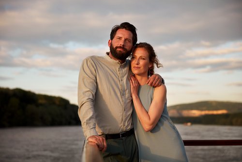 A couple embraces on a boat at dusk with a river and wooded hills in the background.