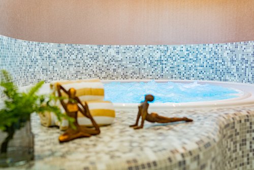 Indoor whirlpool with blue water bubbles, surrounded by mosaic tiles in shades of blue and gray, with two striped towels and two small bronze sculptures in yoga poses in the foreground.