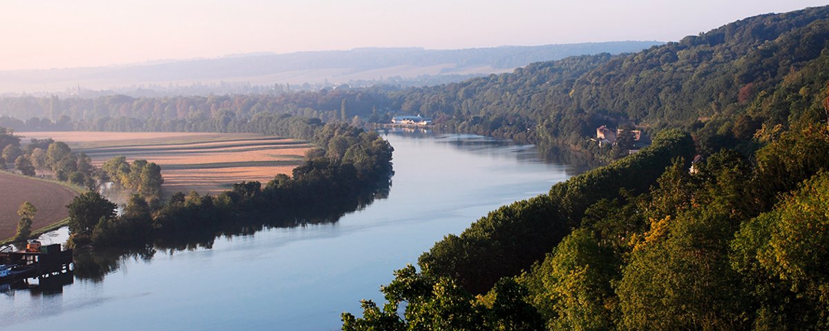 The Seine River flows through wooded hills and fields under a clear sky in Normandy.