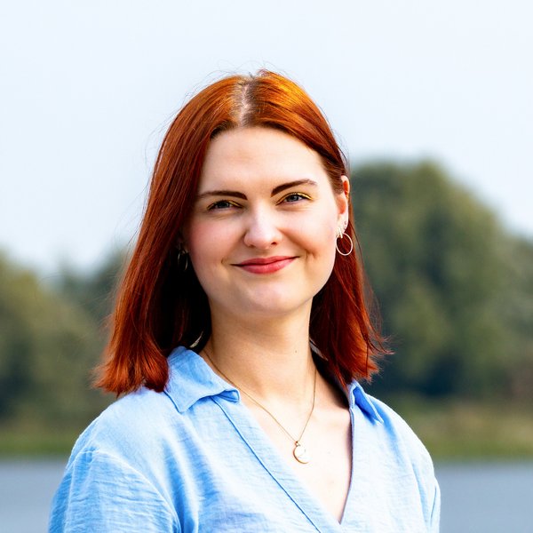 Nele Gaukel with red hair, a blue shirt, and a moon pendant, smiling in front of a lake and trees in the background