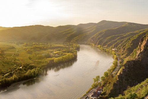 Donautal bei Abendstimmung. Links vom Fluss liegt ein kleines Dorf. Auf der rechnetn Flusseite türmen sich waldbesäumte Hügel, auf denen eine Burgruine liegt.