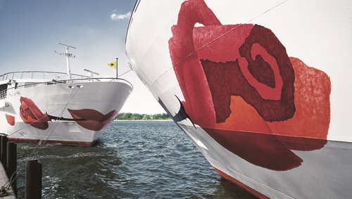 Close-up of a river cruise ship in the A-ROSA fleet with a distinctive rose on the bow in the foreground. Opposite is a second A-ROSA ship with a view of the bow.