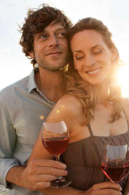 A couple stands at sunset with a glass of wine in hand at the railing of a riverboat. In the background, the river and a riverside landscape.