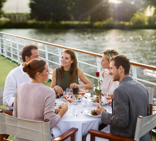 Yvonne Catterfeld and four other people are sitting at a set table on a sun deck by the water, enjoying a meal at sunset.