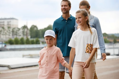A family with two children walking along a riverbank, the children holding hands, with the father and mother following in the background.