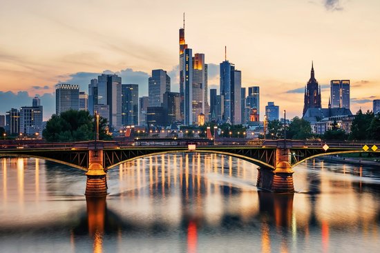 Blick auf die Frankfurter Skyline mit beleuchteten Hochhäusern und einer Brücke über den Fluss im Vordergrund bei Sonnenuntergang