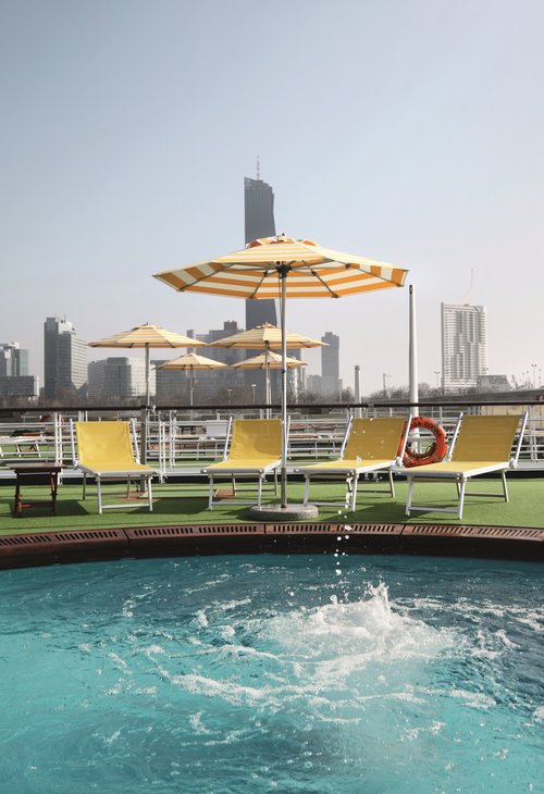Sun deck of the river cruise ship A-ROSA DONNA with a pool in the foreground, surrounded by deck chairs and sun umbrellas, with skyscrapers in the background.