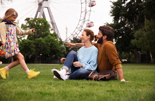 A family enjoys a sunny day in Viennas Prater Park, surrounded by green trees. In the background, there is a Ferris wheel.