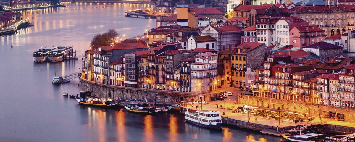 Evening city view of Porto with illuminated buildings on the Douro River and the Arrábida Bridge in the background.