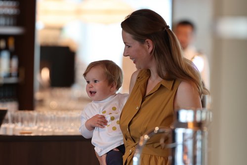 Lachende Frau mit langem hellbraunem Haar und braunem Kleid hält lachendes Kleinkind mit weißem Pullover auf dem Arm.