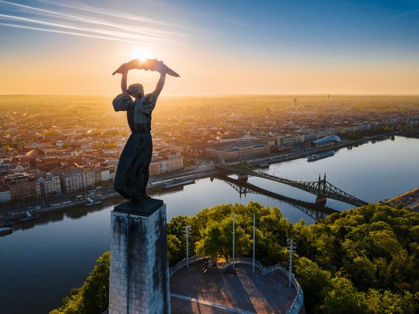 The Statue of Liberty in Budapest on Gellért Hill, surrounded by green trees, overlooking the Liberty Bridge over the Danube and the city in the background at sunset.