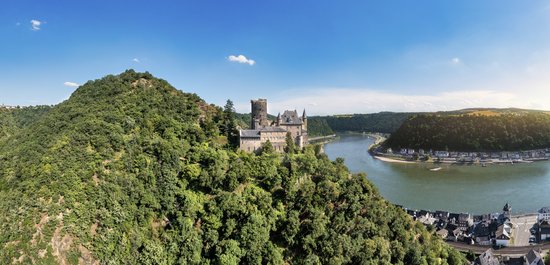Panoramic view of the medieval Katz Castle on a hill in the Middle Rhine, surrounded by vegetated hills and the town of St. Goarshausen on the riverbank.