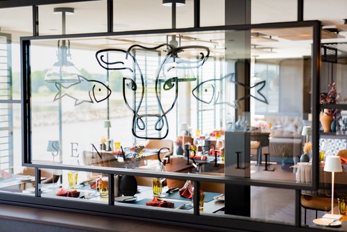 The SENA GRILL restaurant in an industrial-modern design on the A-ROSA SENA with set tables and floor-to-ceiling windows overlooking a tranquil river in daylight.