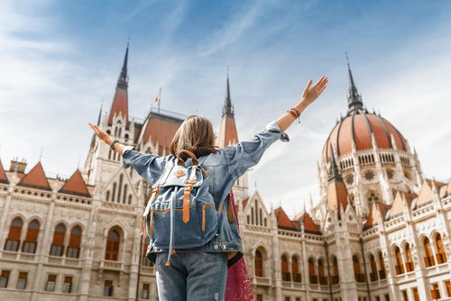 View of a woman with a backpack from behind, stretching her arms upwards and admiring the Hungarian Parliament Building in Budapest in sunny weather.