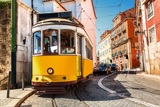 Yellow tram on a narrow cobblestone street in Lisbon, surrounded by colorful buildings and parked cars.