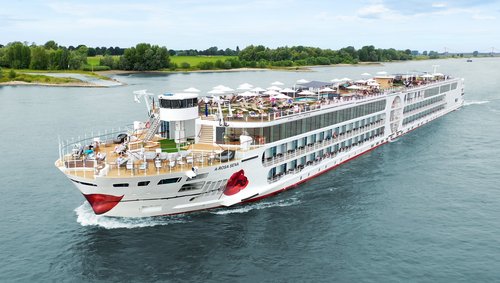 The A-ROSA SENA sails along the Rhine, the area with deck chairs and sunshades is well attended, with the riverbank featuring green landscapes and trees in the background.