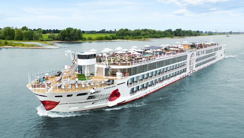 The A-ROSA SENA sails along the Rhine, the area with deck chairs and sunshades is well attended, with the riverbank featuring green landscapes and trees in the background.