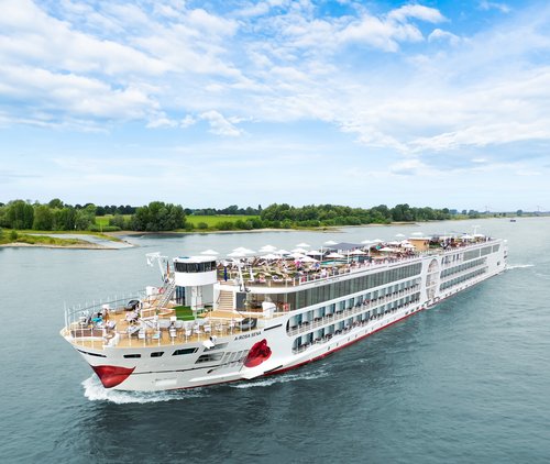 The A-ROSA SENA sails along the Rhine, the area with deck chairs and sunshades is well attended, with the riverbank featuring green landscapes and trees in the background.