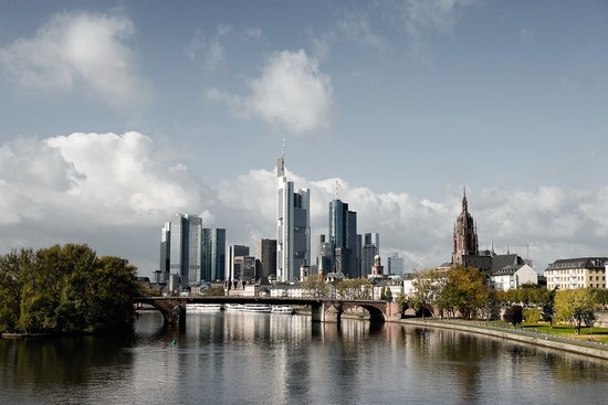 Frankfurt skyline with skyscrapers under a cloudy sky, with the Main River and bridge with park area in the foreground.