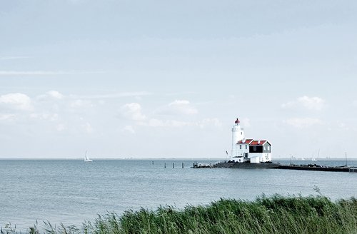 A lighthouse house on the IJsselmeer with reeds in the foreground and sailboats on the sea, under a cloudy sky.