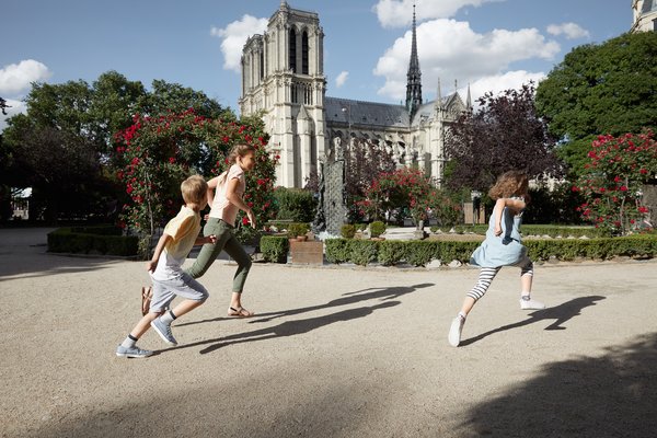 Three children are running in a park with blooming rose bushes in front of the Notre-Dame Cathedral in Paris in sunny weather.