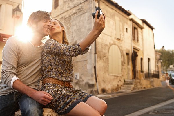A couple sits on a wall in the old town of Arles and takes a picture of themselves with a digital camera. In the background, historical buildings and the setting sun.