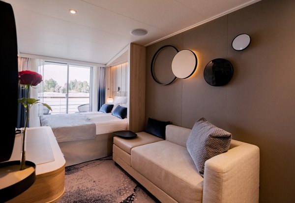 A stylishly furnished cabin on the A-ROSA SENA with a double bed, sofa, floor-to-ceiling windows, and a balcony in daylight.
