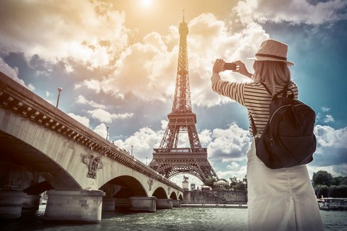 A tourist with a straw hat and backpack photographs the Eiffel Tower under a sunny sky from the Seine bridge in Paris.