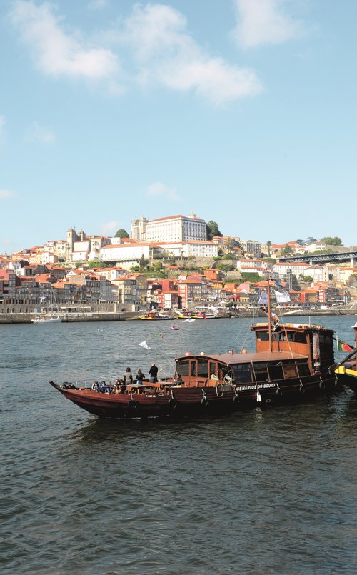 Traditional wooden boat on the Douro River in front of the city of Porto with colorful houses and historic buildings on the shore.