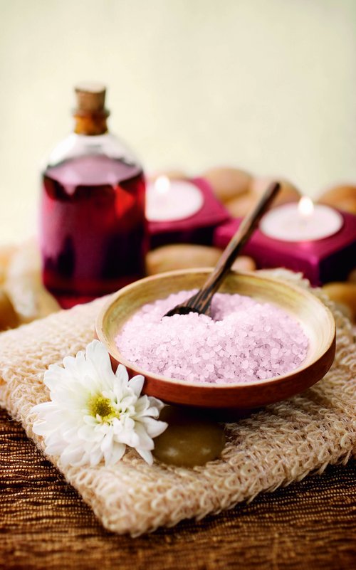 A glass bottle of massage oil and a bowl of salt in a spa setting, adorned with a blossom and candles in the background.
