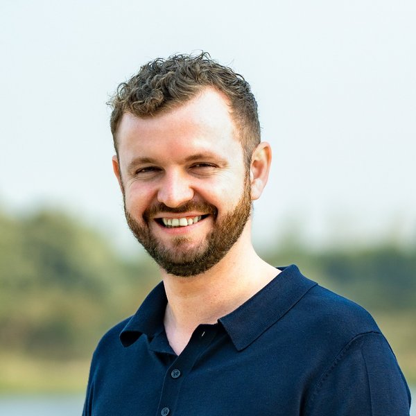 Hauke Heinrich smiles, wears a dark blue polo shirt, stands in front of a blurred natural background with water and trees.