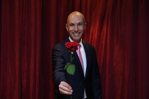 Michael Begasse stands smiling in an elegant suit in front of a curtain and holds a red rose in his hand.
