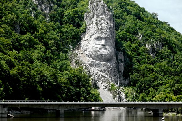 Rocky landscape at the Danube Delta with a rock statue of the head of the Dacian King Decebalus, surrounded by lush vegetation with a bridge and river in the foreground.