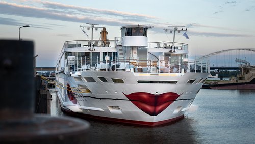 The A-ROSA SENA with a painted kissing mouth and rose on the bow is docked in the evening at a port, the sky is slightly cloudy.