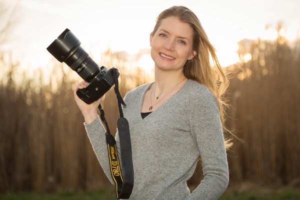 Die Fotografin Lisa Stelzel steht auf einer Wiese mit Ihrer Nikon-Kamera in der Hand und lächelt. Der Hintergrund wird von der untergehenden Sonne erleuchtet.