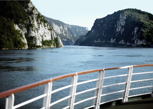 View from a river cruise ship of the vegetated rocky landscape in the Danube Delta. In the foreground is a railing and behind it the quiet river.