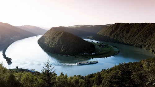 A river cruise ship from the A-ROSA fleet on the Danube along the Schlögener Schlinge. Panoramic view with a look at the adjacent hilly landscape at sunrise.