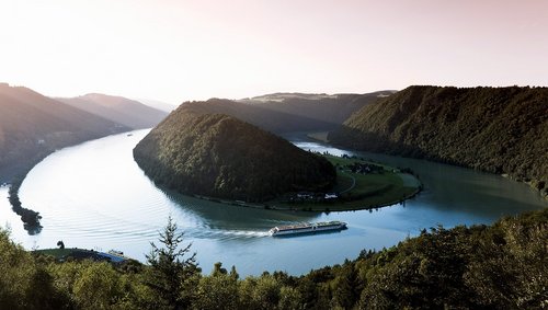 A river cruise ship from the A-ROSA fleet on the Danube along the Schlögener Schlinge. Panoramic view with a look at the adjacent hilly landscape at sunrise.