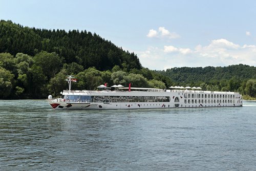 In the afternoon, the A-ROSA SILVA sails along the Rhine past green, wooded hills on the shore; the sky is blue and slightly cloudy.