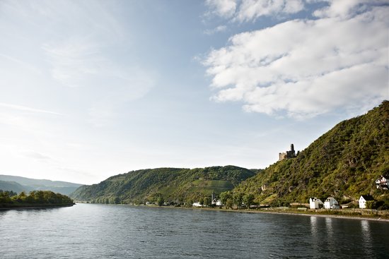 View of the Loreley on the Rhine with the river winding through the landscape, surrounded by green hills with a fortress in the background under a blue, cloudy sky.
