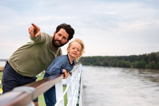 Father and son stand on the deck of a river cruise ship and look over the railing at the river. The father points to something in the distance.