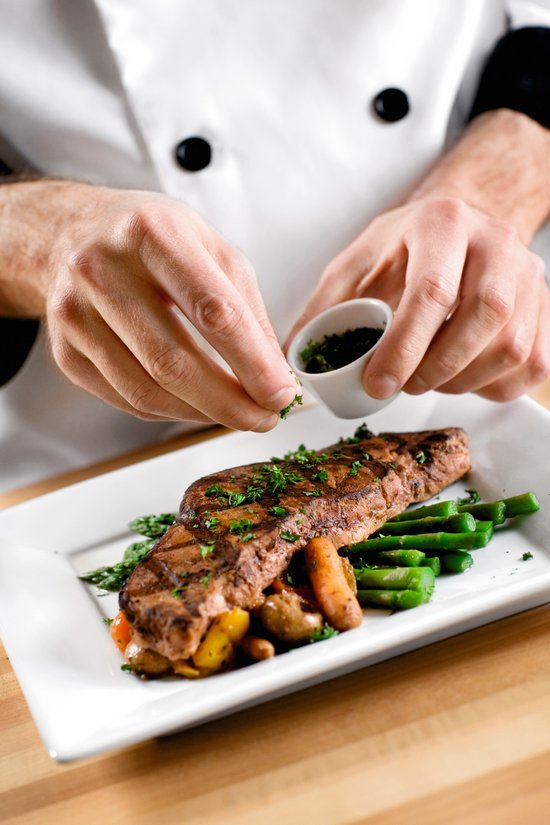 Cook sprinkles fresh herbs on grilled steak with green beans and vegetables on a white plate.