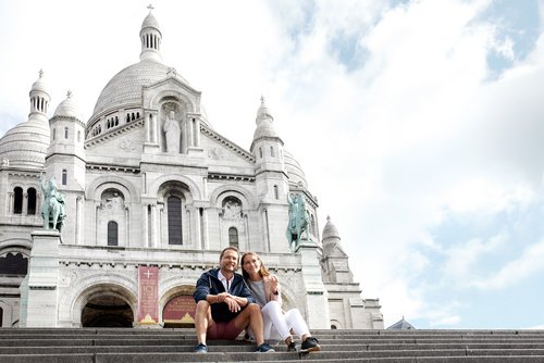 Couple sitting on the steps in front of the Basilica of Sacré-Cœur in Paris under a cloudy sky.