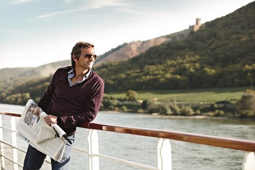 A man with sunglasses is holding a newspaper on the sun deck of a river cruise ship in the sunshine. He is enjoying the view of the hilly landscape along the Rhine.