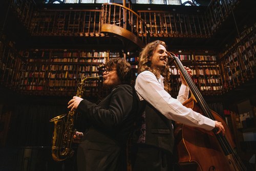 Two musicians are standing back to back in a library during a performance. The person on the left is playing saxophone and the man on the right is playing a double bass.