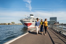 Three people are walking along a pier, with a ship featuring a large red mouth on its bow and a modern building in the background.