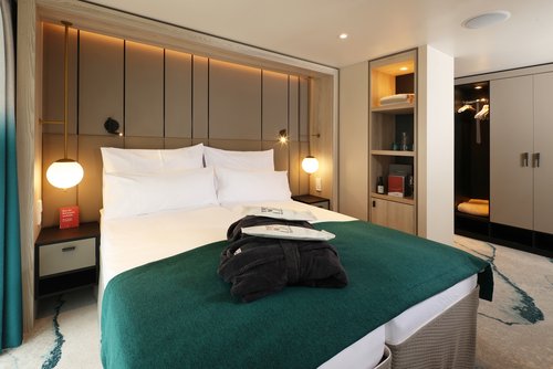 A stylishly furnished suite on the A-ROSA SENA with a double bed, sofa, desk, walk-in closet, floor-to-ceiling windows, and a balcony in daylight.