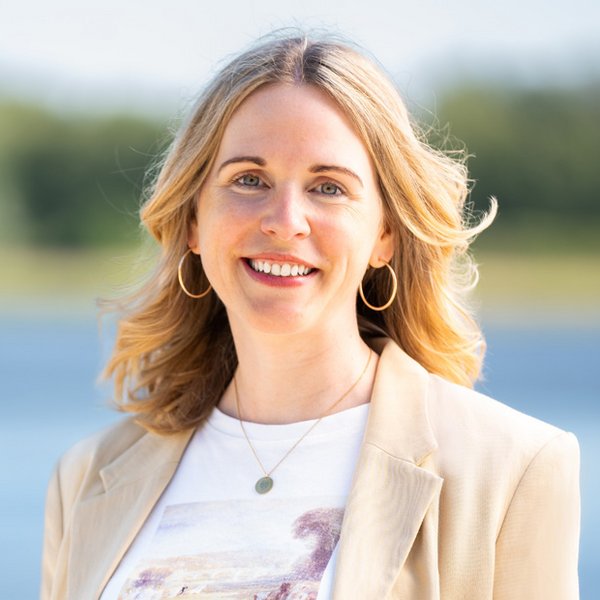 Annika Schmied with blonde hair, beige blazer, white T-shirt, and gold hoop earrings against a blurred background with water