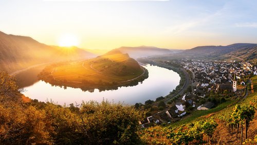A river bend of the Moselle at the Bremm viewpoint overlooking Bremm at sunset, surrounded by wooded hills and vineyards, under a clear sky.