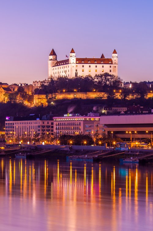 View of the Danube riverbank in Bratislava at sunset with Bratislava Castle in the background, surrounded by trees and the city. In the foreground, the lights are reflected in the river.